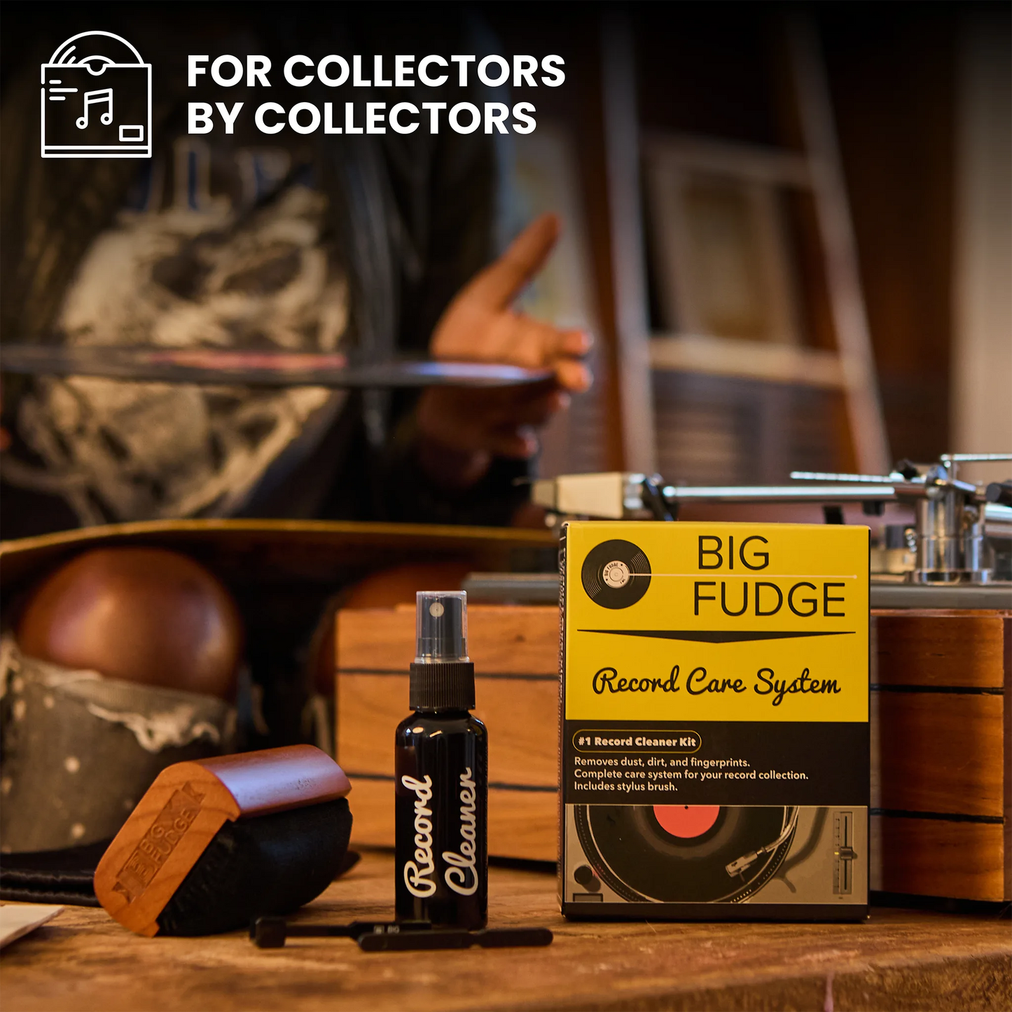 A record care kit displayed on a wooden table in a vintage record shop setting, featuring a yellow and black 'Big Fudge Record Care System' box, a black 'Record Cleaner' spray bottle, a wooden brush, and a turntable in the blurred background. The text 'FOR COLLECTORS BY COLLECTORS' is visible in the top left corner.