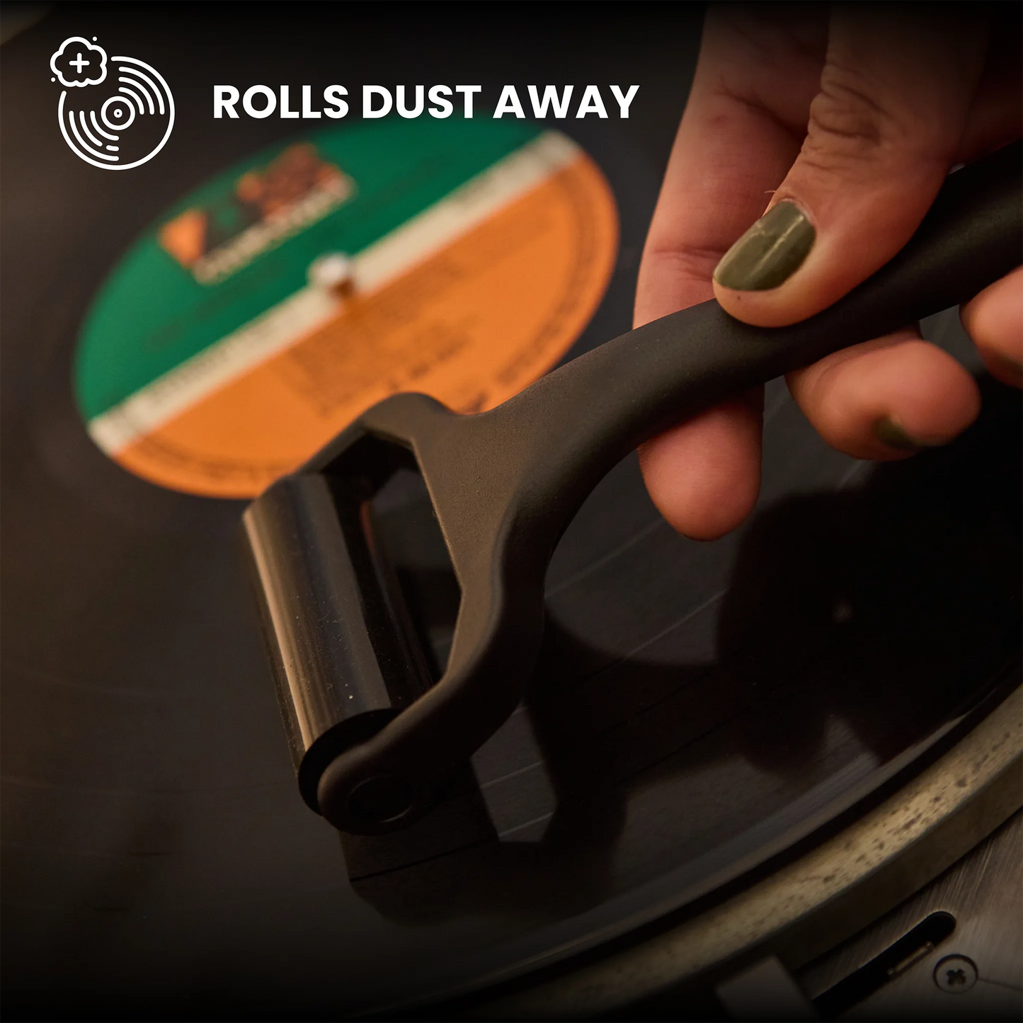 A close-up of a hand holding a black vinyl record cleaning roller over a spinning black record, with the text 'ROLLS DUST AWAY' and a circular logo in the top left.