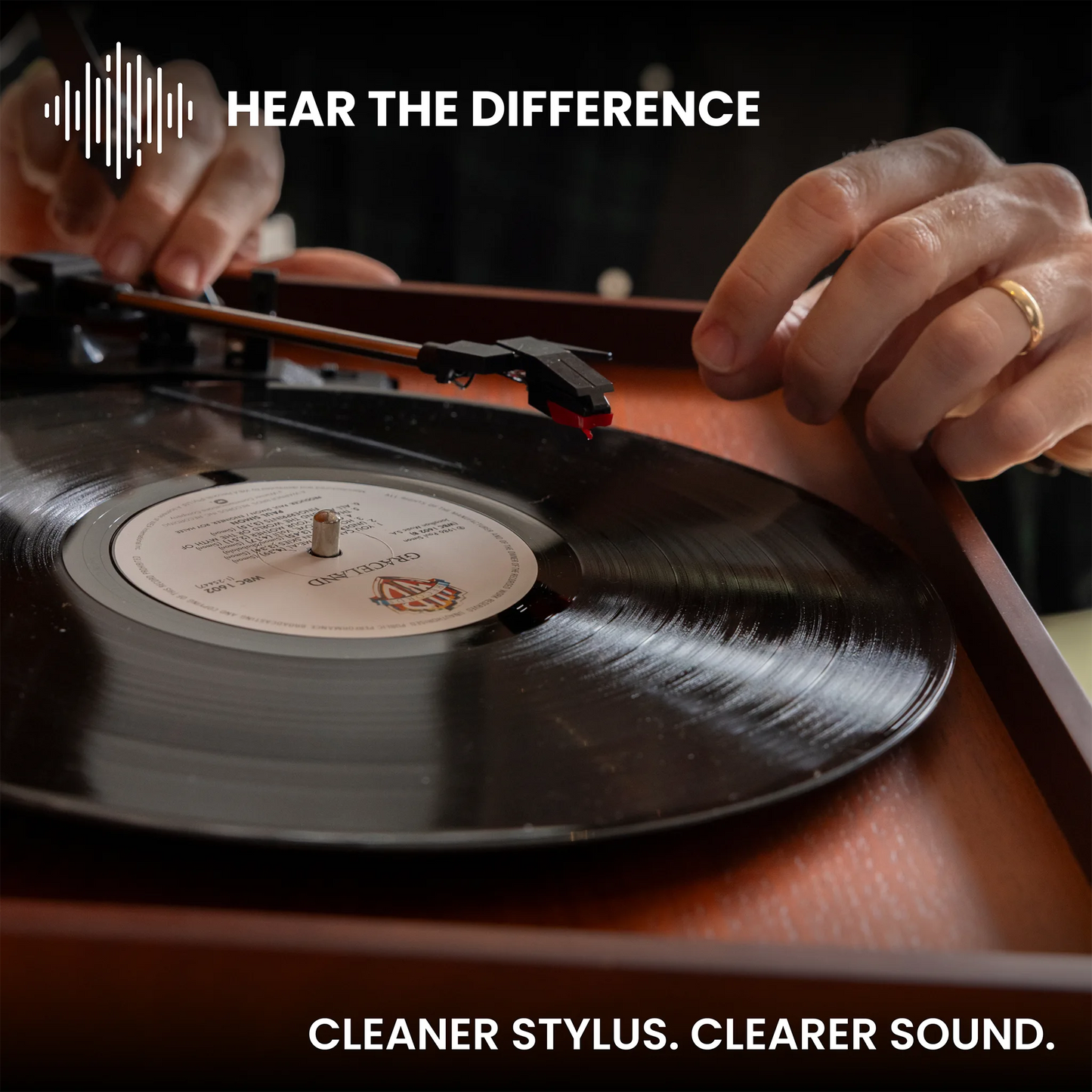 A close-up of a vinyl record on a turntable with a stylus being placed on the tonearm, with hands visible and text reading 'HEAR THE DIFFERENCE' and 'CLEANER STYLUS. CLEARER SOUND.