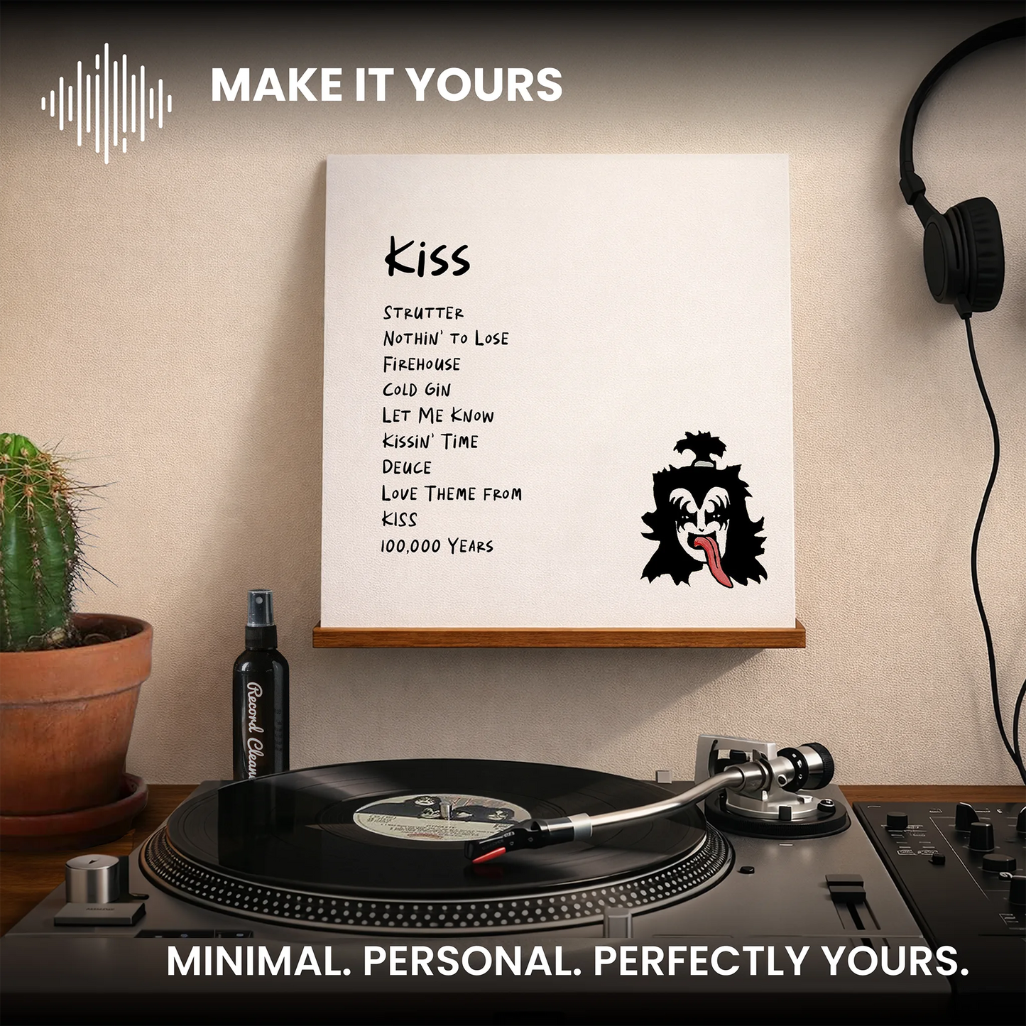 A framed music print featuring the song 'Kiss' with a black-and-white gorilla mask graphic, displayed on a wall above a turntable in a styled room with headphones, a cactus, and a bottle. _Glossy White