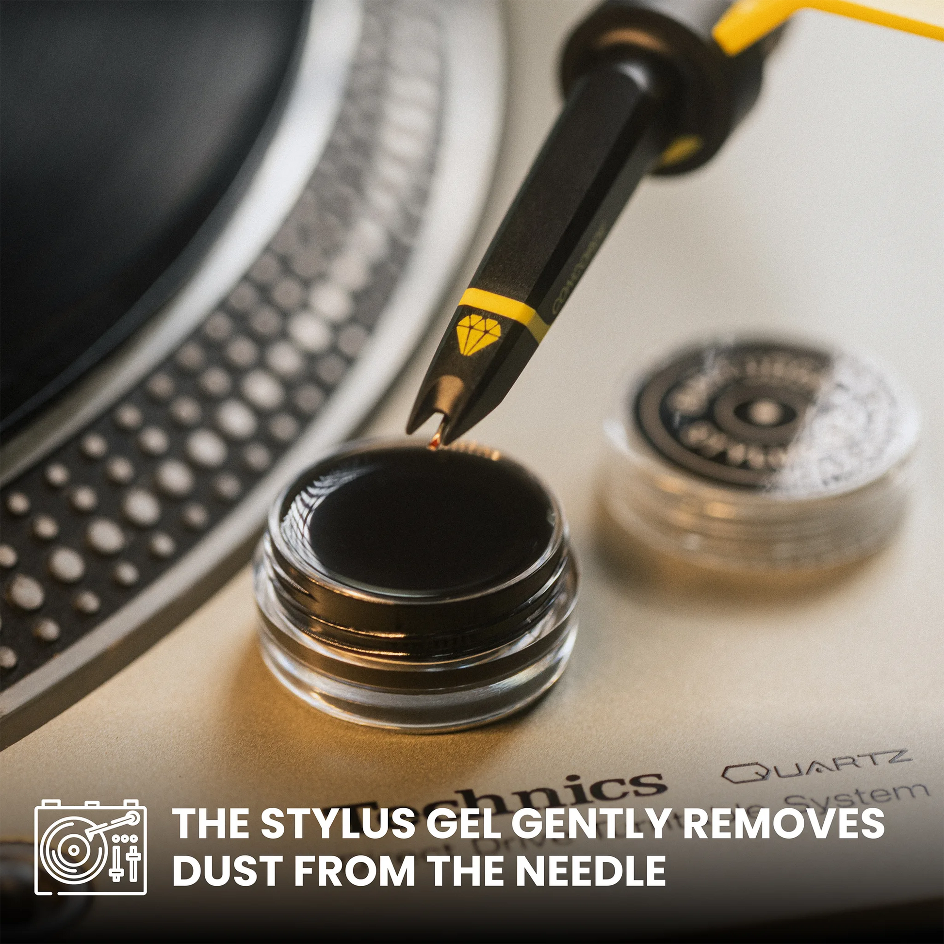 A close-up of a stylus gel pen being used to apply gel to a small clear jar, with the text 'THE STYLUS GEL GENTLY REMOVES DUST FROM THE NEEDLE' overlaid at the bottom. The stylus has a black body with a yellow accent band and a small yellow diamond-like logo, and the background shows part of a turntable with metallic silver and black tones.