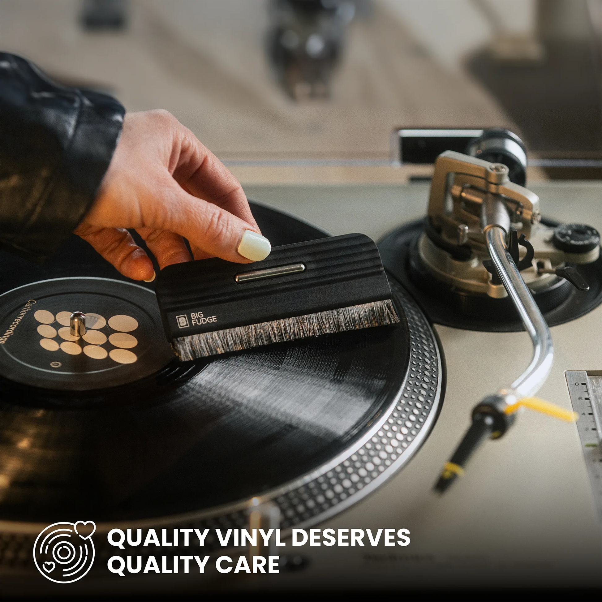 A close-up of a hand using a black vinyl record cleaning brush on a black vinyl record on a turntable, with the text 'QUALITY VINYL DESERVES QUALITY CARE' at the bottom.