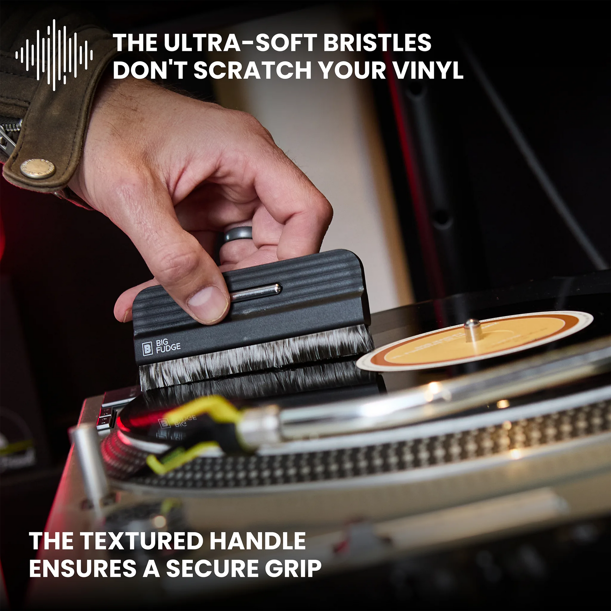 A close-up of a hand using a small black vinyl record cleaning brush on a turntable. Text overlays highlight the ultra-soft bristles and textured handle, with a vinyl record partially visible on the platter.