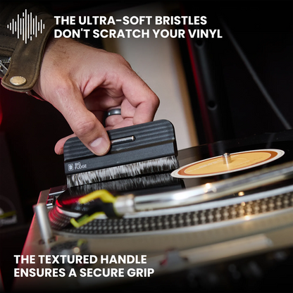 A close-up of a hand using a small black vinyl record cleaning brush on a turntable. Text overlays highlight the ultra-soft bristles and textured handle, with a vinyl record partially visible on the platter.