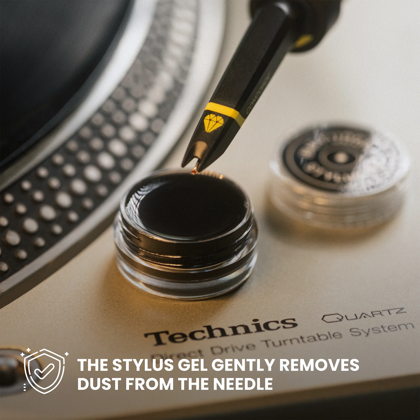Technics stylus gel in a small clear jar, with a stylus tip resting on the gel and a second jar in the background. Text at the bottom reads, 'THE STYLUUS GEL GENTLY REMOVES DUST FROM THE NEEDLE.