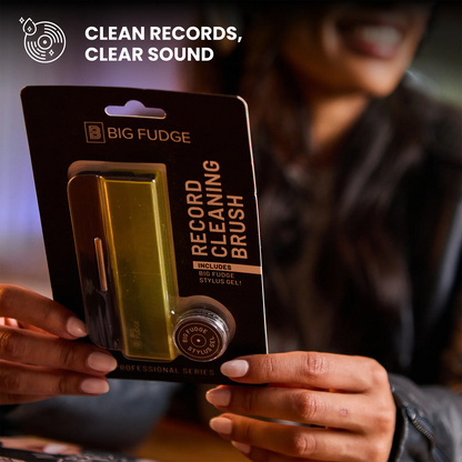 A person holds a packaged Big Fudge record cleaning brush in a dark indoor setting. The product is shown in its retail blister packaging, with the brush visible through a clear window.