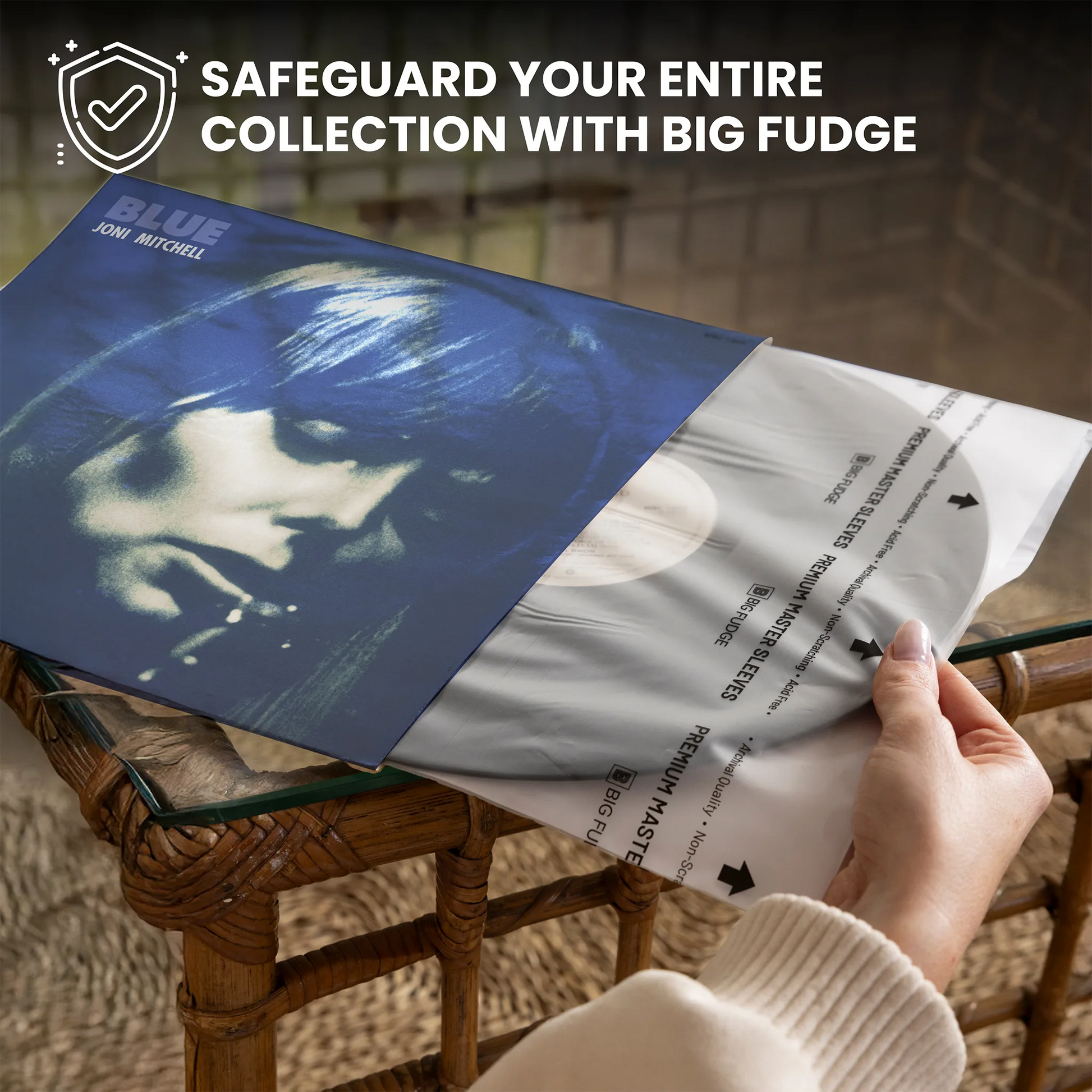 A vinyl record sleeve is being opened by a hand over a rattan table, revealing the inner paper sleeve with track listings. The sleeve features a blue-toned portrait of Jon Jon Jon Jon with the title 'BLUE' and text reading 'SAFEGUARD YOUR ENTIRE COLLECTION WITH BIG FUDGE' at the top.
