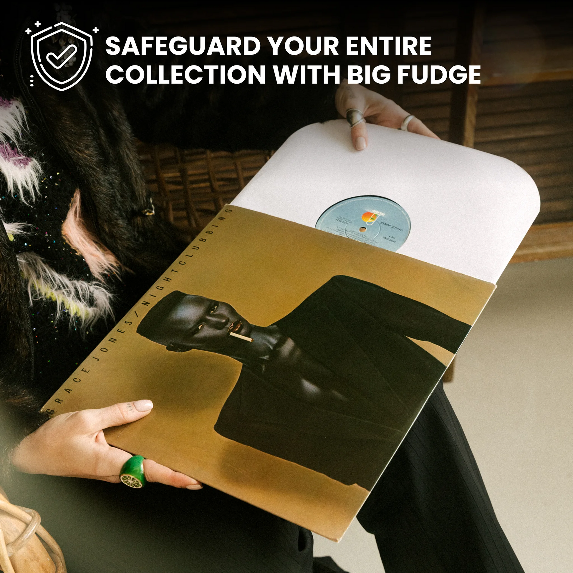 A person holds a vinyl record sleeve featuring a black-and-white portrait of a figure on a gold background, with a white record partially visible. The scene is indoors, and promotional text at the top reads, 'SAFEGUARD YOUR ENTIRE COLLECTION WITH BIG FUDGE. _12 inch