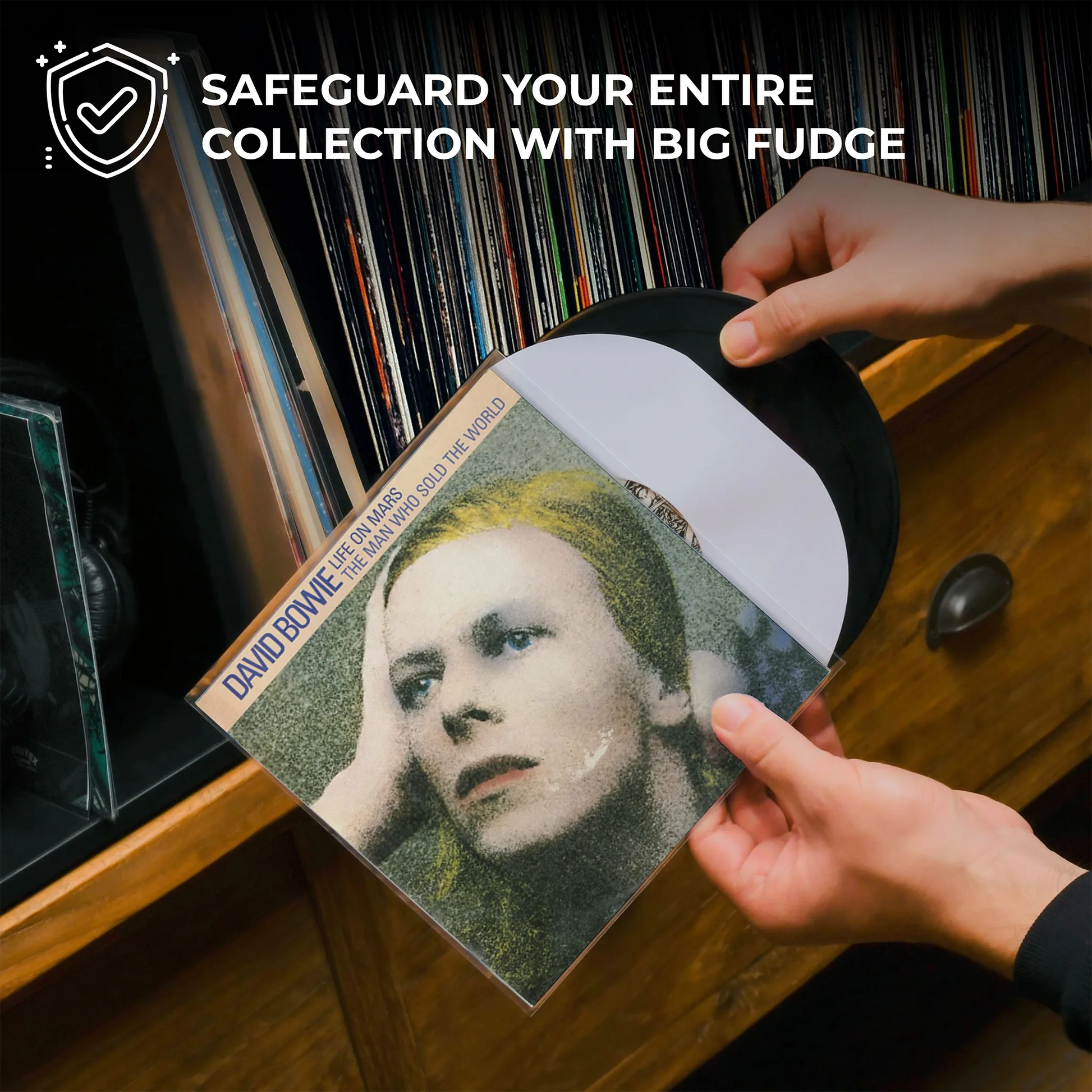 A person's hands are holding a David Bowie 'The World Is Not Enough' vinyl record, showing the black LP and white inner sleeve against a background of a wooden record cabinet filled with vinyl records. A promotional text overlay at the top reads 'SAFEGUARD YOUR ENTIRE COLLECTION WITH BIG FUDGE'. _7 inch